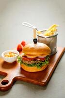 Classic Cajun Beef burger include cheese slice, tomato, onion and lettuce leaf served on wooden board with dip and french fries bucket isolated on grey background side view of appetizer fast food photo