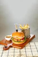 Spicy Naga Beef burger include cheese slice, tomato, onion and lettuce leaf served on wooden board with dip and french fries bucket isolated on grey background side view of appetizer fast food photo