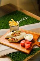 Smoky Chicken Egg Cheese wrap roll filled with tomato, onion, cucumber, with french fries, mayo dip and watermelon slice served on wooden board top view of fastfood appetizer photo