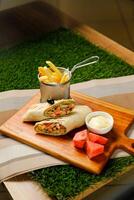 Smoky Chicken Egg Cheese wrap roll filled with tomato, onion, cucumber, with french fries, mayo dip and watermelon slice served on wooden board top view of fastfood appetizer photo