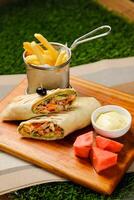 Smoky Chicken Egg Cheese wrap roll filled with tomato, onion, cucumber, with french fries, mayo dip and watermelon slice served on wooden board top view of fastfood appetizer photo