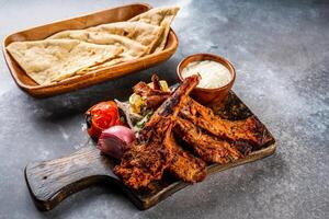 Mutton Champ or lamb chop bbq grill with pita bread, nan, tandoori roti, raita, dip and salad, tomato onion and lemon served in wooden board top view on background spicy barbeque photo