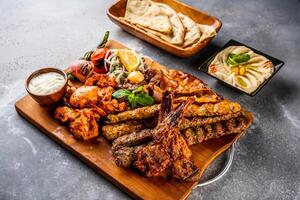 Mix grill with shish tawook, mutton chop, champ, chicken kabab, beef kebab, tikka boti, reshmi and malai, hummus, pita bread, tandoori roti, raita, salad and lemon served in wooden board top view photo