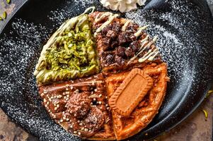 Assorted waffle with Ferrero Rocher, Nutella, Oreo, Lotus, Brownie, Pistachio, Chocolate, Whipped cream, knife and fork served in dish isolated on dark background closeup top view cafe dessert food photo