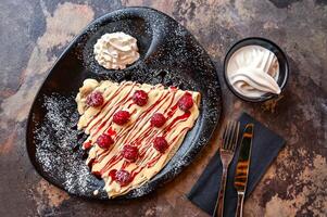 White Chocolate And Raspberry Crepe with whipped cream, knife and fork served in dish isolated on dark background closeup top view of cafe baked dessert food photo