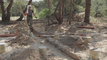 A worker, clad in work attire and equipped with safety gear, is in the process of laying large underground pipes among the partially buried concrete foundations in a forested construction site. video