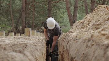 A worker wearing a white hard hat is actively digging a narrow trench in a forested area. The surrounding terrain is uneven with visible tree stumps and soil heaps. video