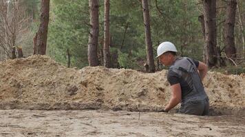 A worker wearing a white hard hat is actively digging a narrow trench in a forested area. The surrounding terrain is uneven with visible tree stumps and soil heaps. video