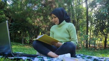 Relaxed muslim woman enjoying weekend at park, sitting on grass and writing book, empty space photo