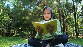 relajado musulmán mujer disfrutando fin de semana a parque, sentado en césped y leyendo libro, vacío espacio foto