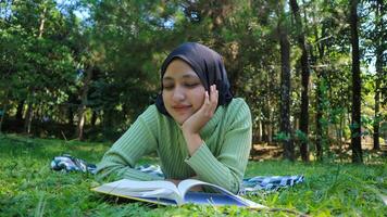 relajado musulmán mujer disfrutando fin de semana a parque, sentado en césped y leyendo libro, vacío espacio foto
