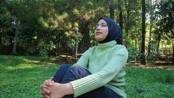 relajado musulmán mujer disfrutando fin de semana en parque sentado en césped, clausura ojos y respiración Fresco aire foto