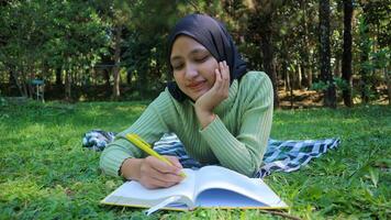 relajado musulmán mujer disfrutando fin de semana a parque, sentado en césped y escritura libro, vacío espacio foto