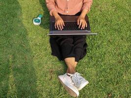 parte superior ver de mujer sentado en parque en el verde césped con computadora portátil, manos en teclado. foto