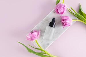 Cosmetic product, oil, serum for face and body skin care in a matte bottle with a dropper lies on a marble podium with delicate tulips. Top view. photo