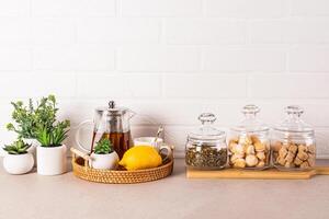 Stylish kitchen background with glass tea pot, lemon, cup, tea jars, sugar, meringue pastry. Green plants in pots. Front view. photo