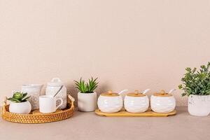 White ceramic tea set on round wicker tray among indoor plants on kitchen countertop. Kitchen background in light colors. A copy space. Front view. photo