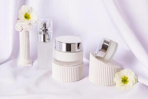 A set of cosmetics in various packages on white podiums, an antique column on a background of white satin. Front view. Product presentation photo