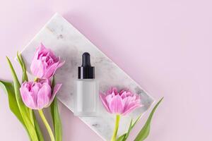 Cosmetic product, oil, serum for face and body skin care in a matte bottle with a dropper lies on a marble podium with delicate tulips. Top view. photo