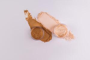 Top view of two glass jars with dry powder in beige and bronze color standing on powder swatches. white background. layout. Product advertising. photo