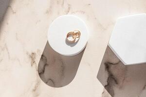 Two gold wedding classic rings lie on a white round podium on a marble background in the rays of the sun. Top view. A copy of the space. mockup. photo