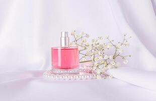 Glass bottle of pink perfume on white satin background with gypsophila branch and pearl beads. The concept of natural floral scents. Front view. photo