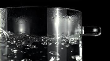 A glass of water is boiling in a pot on a black background, creating steam. The liquid in the highball glass is heating up in the barware video