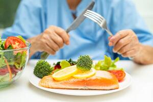 Asian elderly woman patient eating Salmon steak breakfast with vegetable healthy food while sitting and hungry on bed in hospital. photo