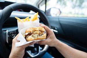 Asian lady holding hamburger and French fries to eat in car, dangerous and risk an accident. photo