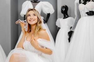 Bride-to-be wearing a wedding dress at a fitting room in a salon, choosing shoes for a wedding and having fun using a shoe as a phone photo