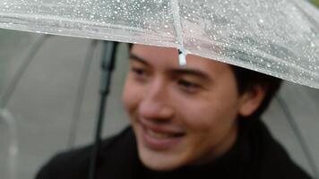 Close-up of a young happy man in a coat who standing under a wet transparent umbrella in the autumn city park and smiling when feeling moments of joy in rainy weather video