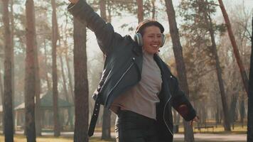 Young happy caucasian girl in headphones and a leather jacket riding on a skateboard with raised hands and smiling in a city park on a sunny day. Concept of interesting autumn urban leisure video