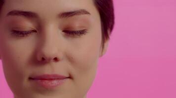 retrato de un joven atractivo mujer apertura su ojos, mirando a el cámara y sonriente con un despreocupado positivo cara en un rosado antecedentes en el estudio cerca arriba video