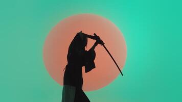 Silhouette of a young asian woman in a traditional kimono slowly raising the katana over her head in the studio with a turquoise blue green background and red sun video