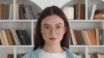 porträtt av en ung leende caucasian kvinna i glasögon höjning henne huvud och ser på de kamera med de bakgrund av vit bokhyllor med många böcker på de bokhyllor video