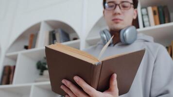 Young asian man in glasses and headphones close up holding a book, flipping its pages while getting his education with the bookcases in the backdrop video