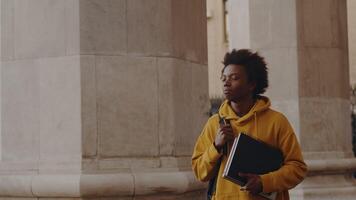 Young black programmer walking alongside the building outdoors after a successful working day with a smiling satisfied face holding a laptop and copybook video