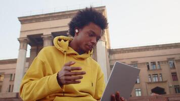 Young black guy in casual clothes talking with a friend on a call with a laptop and wireless headphones with a building in a backdrop, close up, bottom view video