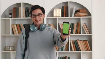 giovane asiatico uomo con cuffie e un' zaino mostrando un' smartphone con un' modello verde schermo e sorridente con il sfondo di bianca librerie con molti libri su il libreria video