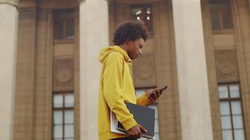 Young black programmer walking outdoors with a laptop and copybook and scrolling messages from a client on his smartphone with a building on a backdrop video