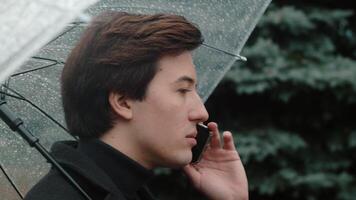 Young man in a coat close up walking under a transparent umbrella in the autumn city park and talking on a smartphone with an enthusiastic face video