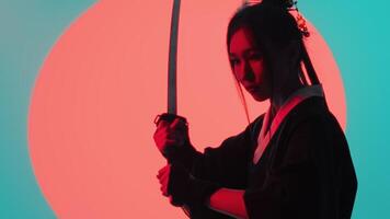 Young asian woman in a traditional kimono close up holding a raised katana with both hands and standing still in the studio with a turquoise blue green background and red sun video