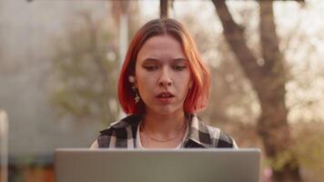 Young girl with red hair wearing casual clothes close up working on a laptop outdoors, getting upset and covering her face with her hand when finding out about a serious error in a working process video