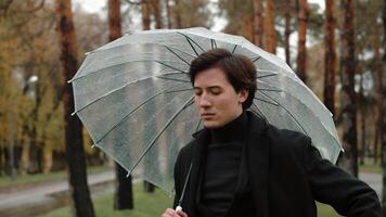 Jeune homme dans une manteau permanent en dessous de une transparent parapluie dans un l'automne ville parc et soufflant le sien nez dans une mouchoir car de humide et du froid temps video