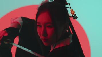 Young asian woman in a traditional kimono close up showing katana control skills with a confident face in the studio with a turquoise blue green background and red sun video