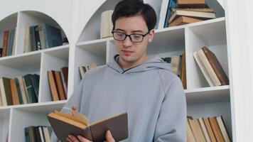 joven asiático hombre en pie con el libro, voltear sus paginas y finalmente hallazgo el necesario información mientras consiguiendo su educación con el librerías en el fondo video