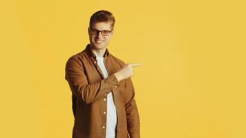 Young smiling caucasian man in glasses and casual clothes pointing persistently with a finger at the side on a yellow background in the studio video