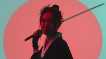 Portrait of a young asian woman in a traditional kimono close up resting the katana on her shoulder in the studio with a turquoise blue green background and red sun video