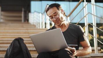 Young asian man in glasses sitting on the stairs with a laptop and talking with a client on a call with a smile on his face. Concept of modern remote work with a flexible schedule video