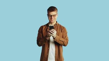 Young attractive caucasian man in glasses and casual clothes typing text on a smartphone, when looking to the camera and smiling on a blue background in the studio video